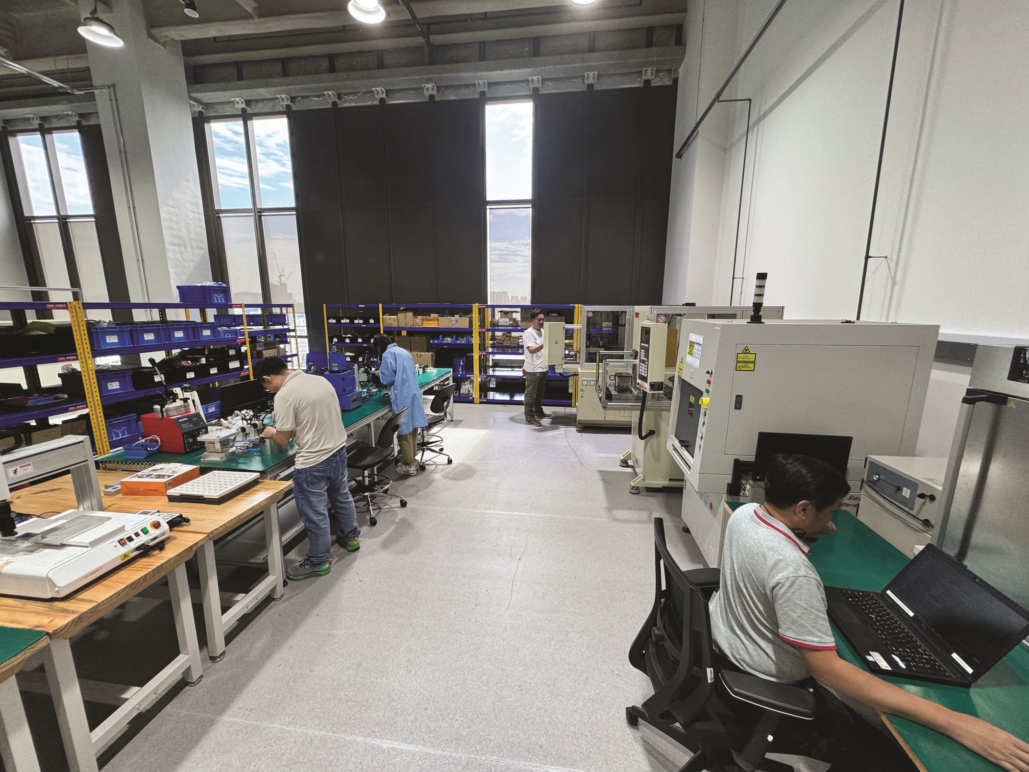 Inside a modern electronics factory, technicians work at benches with equipment, shelving and machinery. Bright windows are in the background.